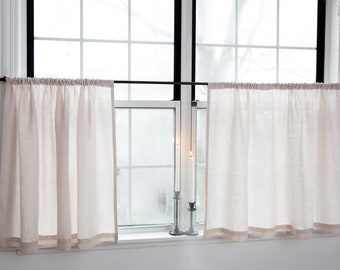 May include: Two beige linen cafe curtains with a light brown border hang on a black rod in front of a window. Two lit candles in silver candlesticks sit on the windowsill. The curtains are gathered at the top.