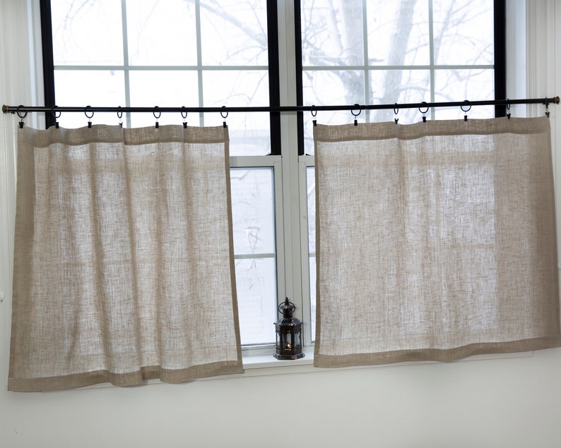 Puede incluir: Dos cortinas de lino beige colgadas de una barra de cortina negra delante de una ventana. Las cortinas est&aacute;n fruncidas en la parte superior y tienen un aspecto sencillo y casual.