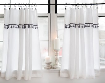 May include: Two white linen curtains with gray tassel trim hang from a black curtain rod. The curtains are hung in front of a window with white trim.