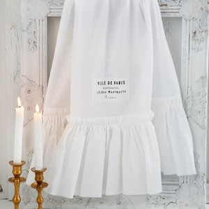 May include: White cotton fabric with a ruffled edge and a black label that reads "Ville de Paris, Nouveautes, 170, Rue Montmartre". The fabric is draped over a white ornate picture frame and two gold candlesticks with white candles.