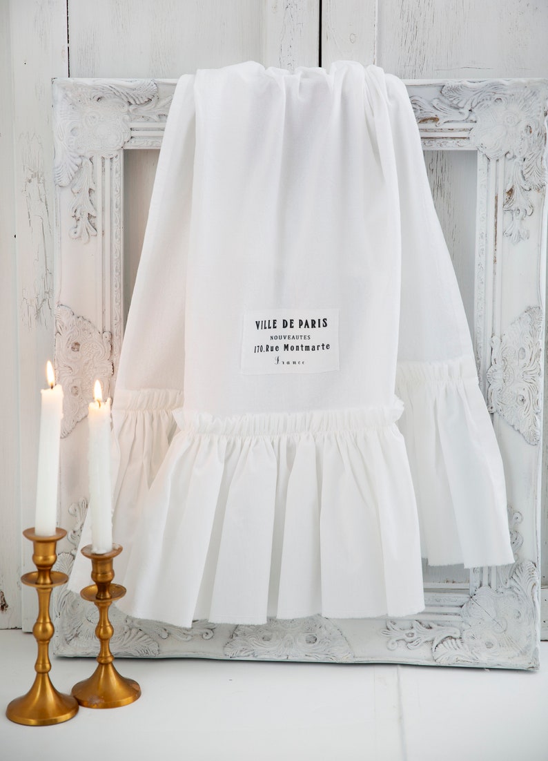 May include: White cotton fabric with a ruffled edge and a black label that reads "Ville de Paris, Nouveautes, 170, Rue Montmartre". The fabric is draped over a white ornate picture frame and two gold candlesticks with white candles.