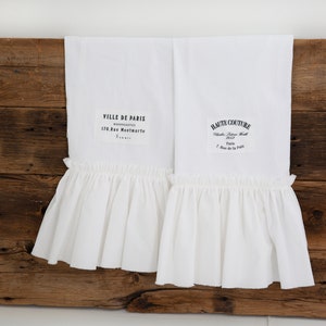 May include: Two white kitchen towels with ruffled edges and black text. The text on the left towel reads "VILLE DE PARIS NOUVEAUTES 170 Rue Montmartre Paris". The text on the right towel reads "HAUTE COUTURE  Chauss.  Femme  H.  1933 Paris 7 Rue de la Paix".