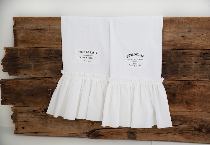 May include: Two white kitchen towels with ruffled edges and black text. The text on the left towel reads "VILLE DE PARIS NOUVEAUTES 170 Rue Montmartre Paris". The text on the right towel reads "HAUTE COUTURE  Chauss.  Femme  H.  1933 Paris 7 Rue de la Paix".