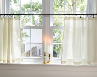 May include: Two white linen curtains hang from a black curtain rod in front of a window with two gold candle holders and white candles on the window sill.