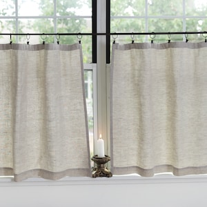 May include: A pair of beige linen cafe curtains with a black metal rod and rings. The curtains are hung on a window with a white frame. A white candle sits on a decorative stand in front of the window.