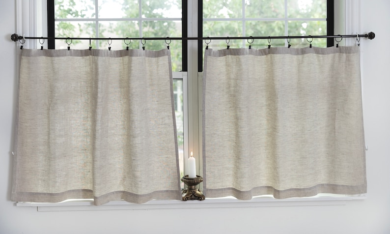 May include: A pair of beige linen cafe curtains with a black metal rod and rings. The curtains are hung on a window with a white frame. A white candle sits on a decorative stand in front of the window.