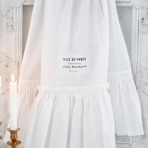 May include: White cotton fabric with a ruffled edge and a black label that reads "VILLE DE PARIS NOUVEAUTES 170, rue Montmartre PARIS".