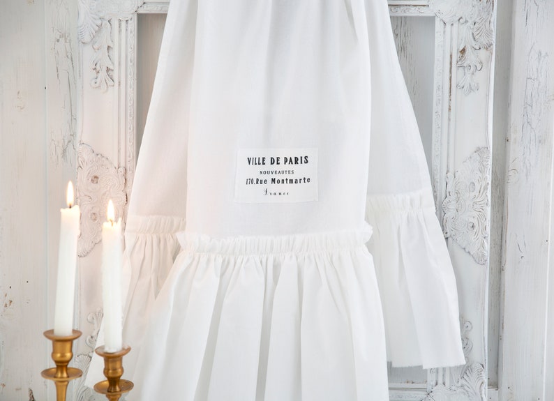 May include: White cotton fabric with a ruffled edge and a black label that reads "VILLE DE PARIS NOUVEAUTES 170, rue Montmartre PARIS".