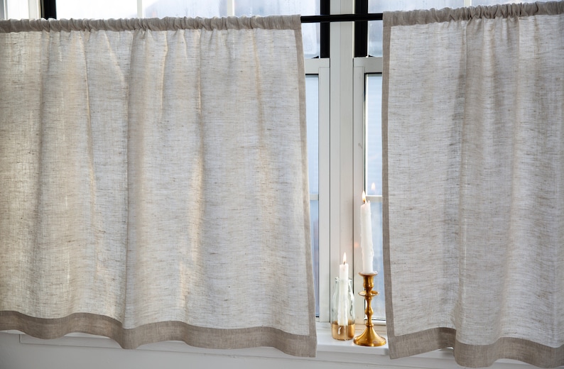 May include: Two white linen cafe curtains with a brown trim hang in a window. Three candles are on the windowsill.