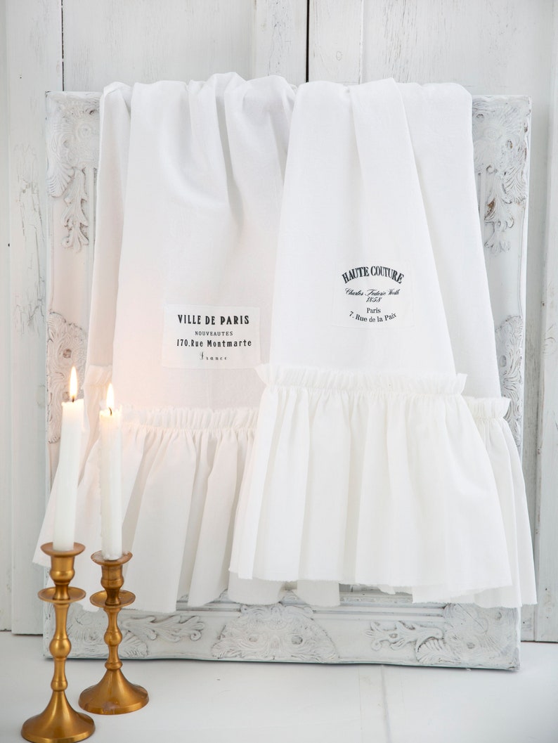 May include: Two white cotton tea towels with ruffled edges. One towel has the text "VILLE DE PARIS NOUVEAUTES 170, Rue Montmartre" and the other towel has the text "HAUTE COUTURE Charles Frederick Worth 1858 Paris 7, Rue de la Paix".