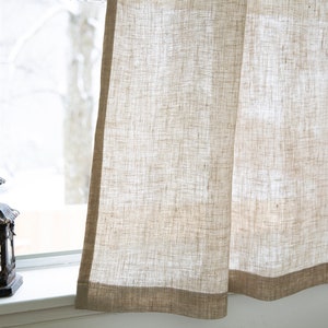 Puede incluir: Un par de cortinas de lino beige con un tejido texturizado sutil. Las cortinas tienen un ribete marr&oacute;n a lo largo del borde inferior.