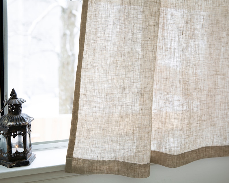 Puede incluir: Un par de cortinas de lino beige con un tejido texturizado sutil. Las cortinas tienen un ribete marr&oacute;n a lo largo del borde inferior.