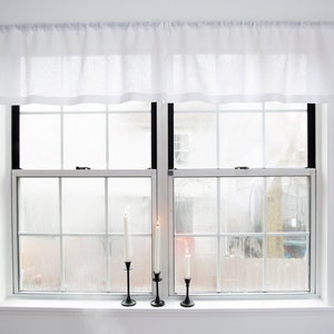 May include: White sheer valance curtains hang above a window with black trim. Three black candle holders with white candles are on the window sill.