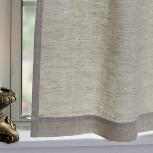 May include: A white candle burning in a gold metal candlestick on a windowsill with a sheer linen curtain with a gray border.