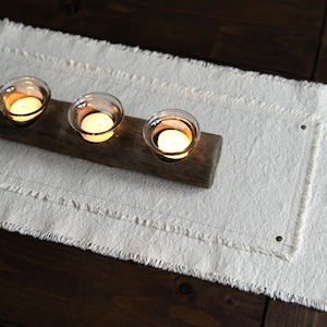 May include: A white table runner with frayed edges and a wooden candle holder with three glass candle holders. The candle holder is placed on the table runner.