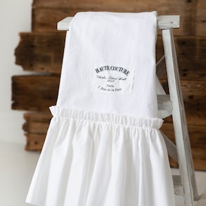 May include: White cotton fabric with a ruffled edge. The fabric has a black label with the text "HAUTE COUTURE Charles Frederick Worth 1858 Paris 7 Rue de la Paix".