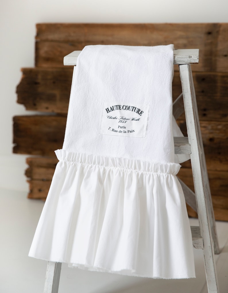 May include: White cotton fabric with a ruffled edge. The fabric has a black label with the text "HAUTE COUTURE Charles Frederick Worth 1858 Paris 7 Rue de la Paix".