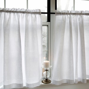 May include: Two white sheer cafe curtains hang from a black curtain rod in front of a window. A white candle sits on a decorative stand in front of the window.
