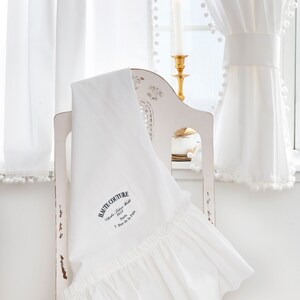 May include: White ruffled fabric with the text "HAUTE COUTURE" embroidered on it. The fabric is draped over a white chair with a gold candle holder in the background.