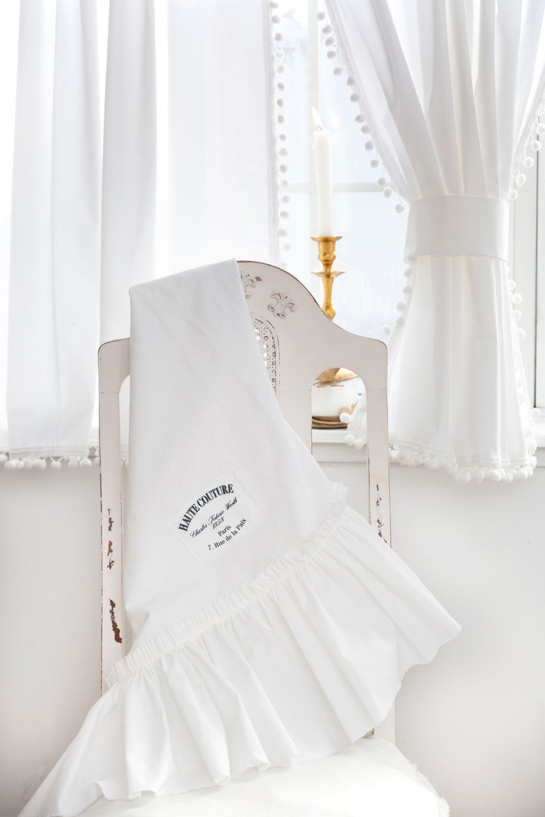 May include: White ruffled fabric with the text "HAUTE COUTURE" embroidered on it. The fabric is draped over a white chair with a gold candle holder in the background.