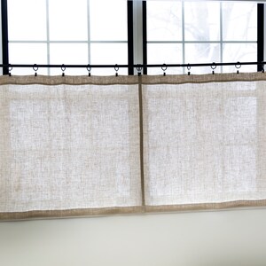 Puede incluir: Un par de cortinas de lino beige colgadas de una barra de cortina negra. Las cortinas son simples y sin adornos, con una textura sutil.