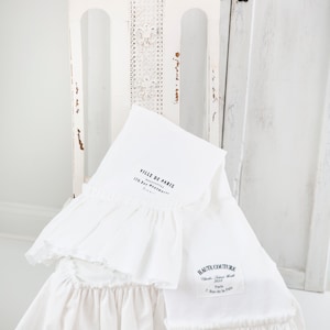 May include: Two white cotton tea towels with ruffled edges. The top towel has the text "Ville de Paris" and "Souvenirs" with the address "170 Rue Montmartre, Paris". The bottom towel has the text "Haute Couture" with the address "Chambre Syndicale de la Haute Couture, Paris, 7 Rue de la Paix".