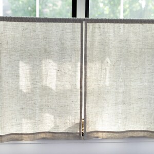 May include: Two white linen curtains with a light gray trim hang in a window. The curtains are slightly translucent, allowing light to shine through.