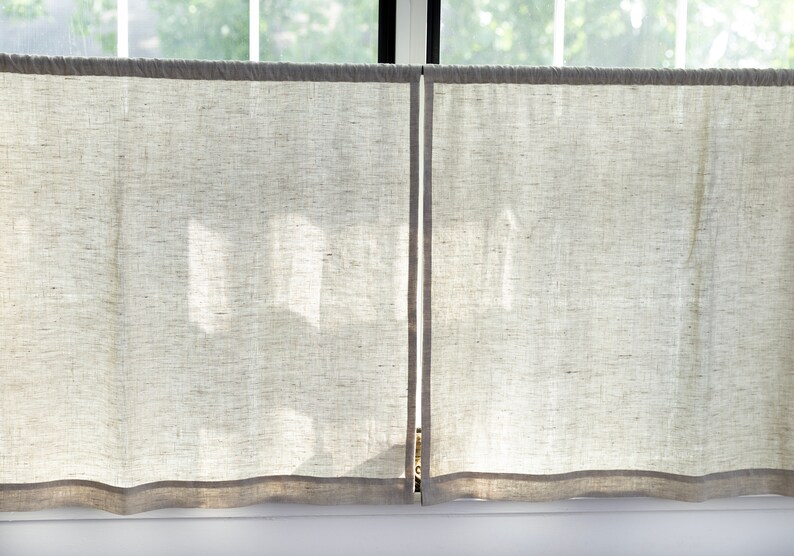May include: Two white linen curtains with a light gray trim hang in a window. The curtains are slightly translucent, allowing light to shine through.
