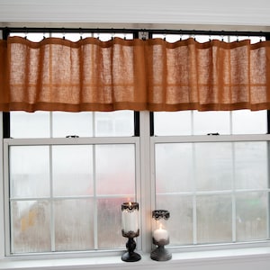 May include: A window with two panes of glass, each with a white frame and black trim. The window is covered with a brown burlap valance with a black rod. Two black metal candle holders with white candles are on the window sill.