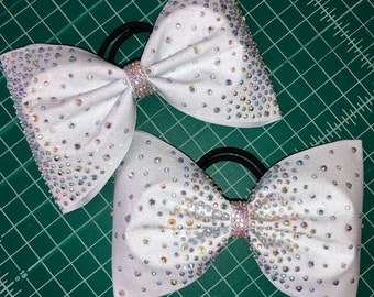 May include: Two white hair bows with sparkling rhinestones. Each bow is adorned with iridescent crystals and a rhinestone-studded center. The bows are attached to black elastic bands, perfect for cheerleading or dance.
