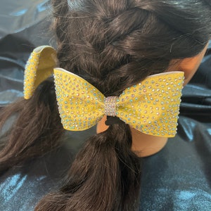 May include: A yellow sparkly hair bow with rhinestones. The bow is tied in a double loop and has a black elastic band in the center.