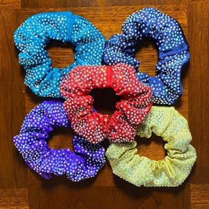 May include: Five colorful hair scrunchies with rhinestones. The scrunchies are in shades of blue, red, purple, yellow, and teal.
