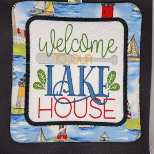 Puede incluir: Un salvamanteles cuadrado acolchado con un fondo de escena de lago azul y blanco. El centro presenta las palabras "Welcome to our Lake House" en bordado verde, azul y rojo. Se adjunta un pequeño lazo azul para colgar.