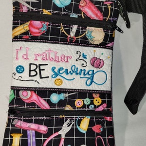 May include: Black fabric crossbody bag with a zippered closure and a black strap. The bag has a white panel with the embroidered text "I'd rather be sewing". The bag is decorated with a sewing themed print.