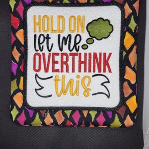 May include: A square, quilted potholder with the text "HOLD ON let me OVERTHINK this" in yellow, red, and black. The potholder has a white center and a black border with colorful geometric shapes.