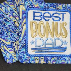 May include: Two blue and gold marbled fabric coasters with a white square patch in the center. The patch has blue embroidered text that reads "Best Bonus Dad" with a gold star on each side.
