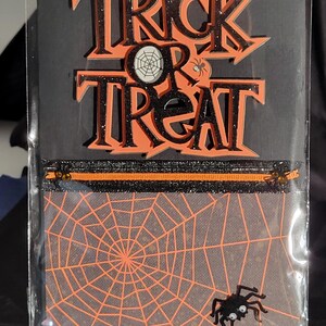 May include: Halloween-themed pouch with the words "Trick or Treat" in orange and black glitter. The pouch features a spider web design and a small spider. The top has a black zipper with orange accents.