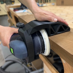 May include: A black sanding jig with a white sanding pad attached to a power sander. The jig is being used to sand a piece of wood.