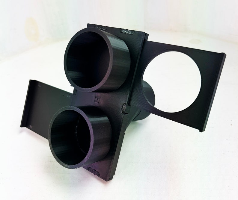 May include: A black 3D-printed object with two cylindrical openings and a circular opening. The object has a rectangular base and appears to be a holder or mount. The design is angular and geometric.