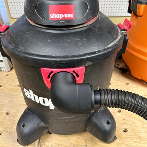 Puede incluir: Una aspiradora Shop-Vac h&uacute;meda/seca negra con una tapa roja y una manguera negra conectada al lateral. La aspiradora est&aacute; sobre una superficie de madera con agujeros.