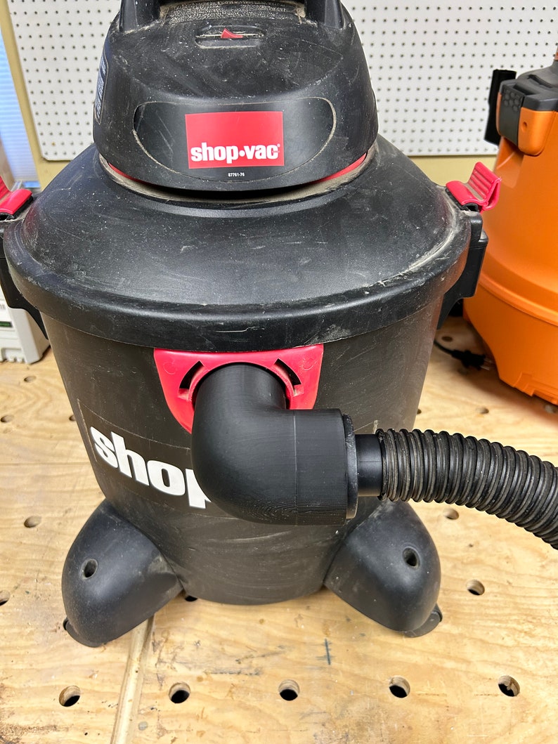 Puede incluir: Una aspiradora Shop-Vac h&uacute;meda/seca negra con una tapa roja y una manguera negra conectada al lateral. La aspiradora est&aacute; sobre una superficie de madera con agujeros.