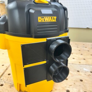 May include: A yellow and black DEWALT wet/dry vacuum with a black attachment featuring two circular openings. The DEWALT logo is prominently displayed in yellow on the front of the vacuum.