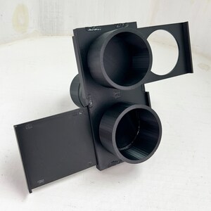 May include: A black 3D-printed object with multiple cylindrical openings and rectangular panels. The object has a complex geometric design, with two large circular openings and a smaller circular opening. The panels extend outward, creating a multi-faceted structure.