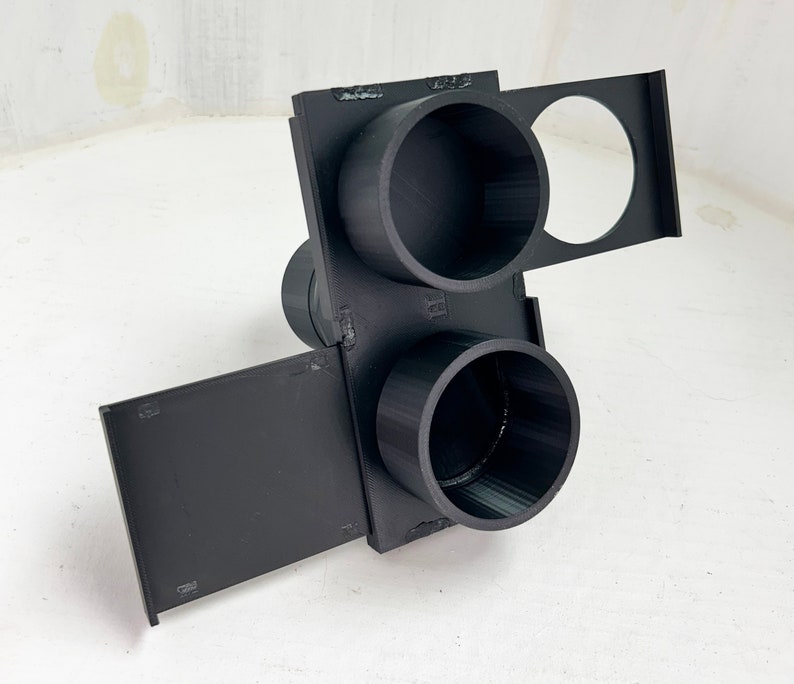 May include: A black 3D-printed object with multiple cylindrical openings and rectangular panels. The object has a complex geometric design, with two large circular openings and a smaller circular opening. The panels extend outward, creating a multi-faceted structure.