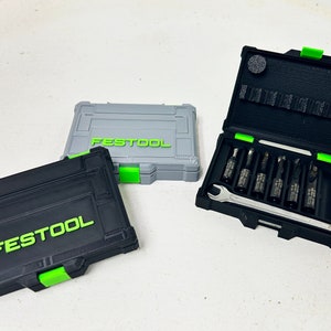 May include: Three black and gray toolboxes with the word "FESTOOL" printed on the front. The open toolbox contains a set of drill bits and a wrench.