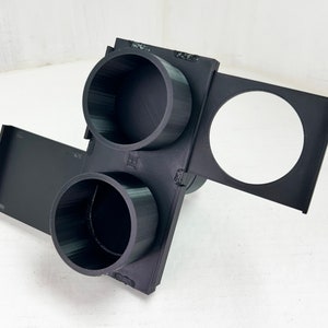 May include: A black 3D-printed object with two cylindrical cup holders and a circular opening. The object has a rectangular base and a hinged section. The design appears to be for holding cups or small items.