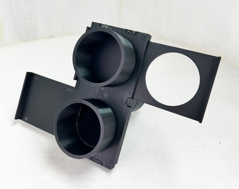 May include: A black 3D-printed object with two cylindrical cup holders and a circular opening. The object has a rectangular base and a hinged section. The design appears to be for holding cups or small items.