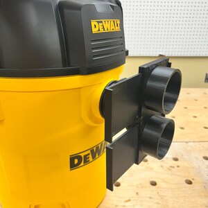 May include: A yellow and black DeWalt wet/dry vacuum with a black attachment. The vacuum has the DeWalt logo in yellow. The attachment has two black circular openings. The vacuum is on a wooden surface.