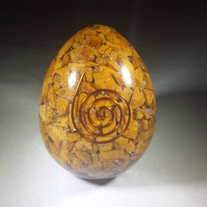 Puede incluir: Un huevo de cristal amarillo dorado con una espiral de alambre de cobre en su interior. El huevo está hecho de pequeños trozos de cristal que se mantienen unidos por una resina transparente.