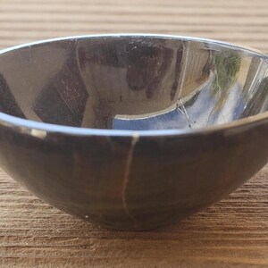 May include: A small, dark gray bowl with a rounded shape. The bowl has a smooth, reflective surface and appears to be made of stone or a similar material. The bowl is sitting on a wooden surface.